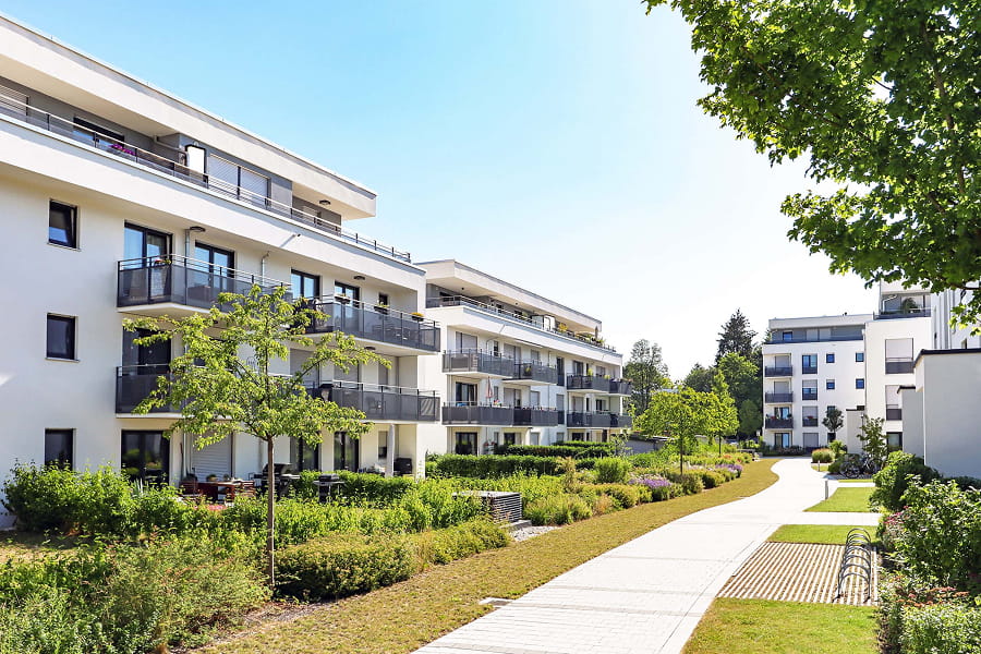 Weiße Mehrfamilienhäuser mit Balkonen, gepflegten Gärten und hellem Fußweg in einer modernen Wohnanlage